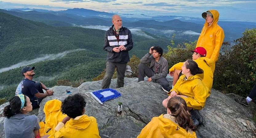 A group of people stand on a rocky overlook, high above the blue ridge mountains. The group is listening to an instructor and many students are wearing yellow rain suits. 